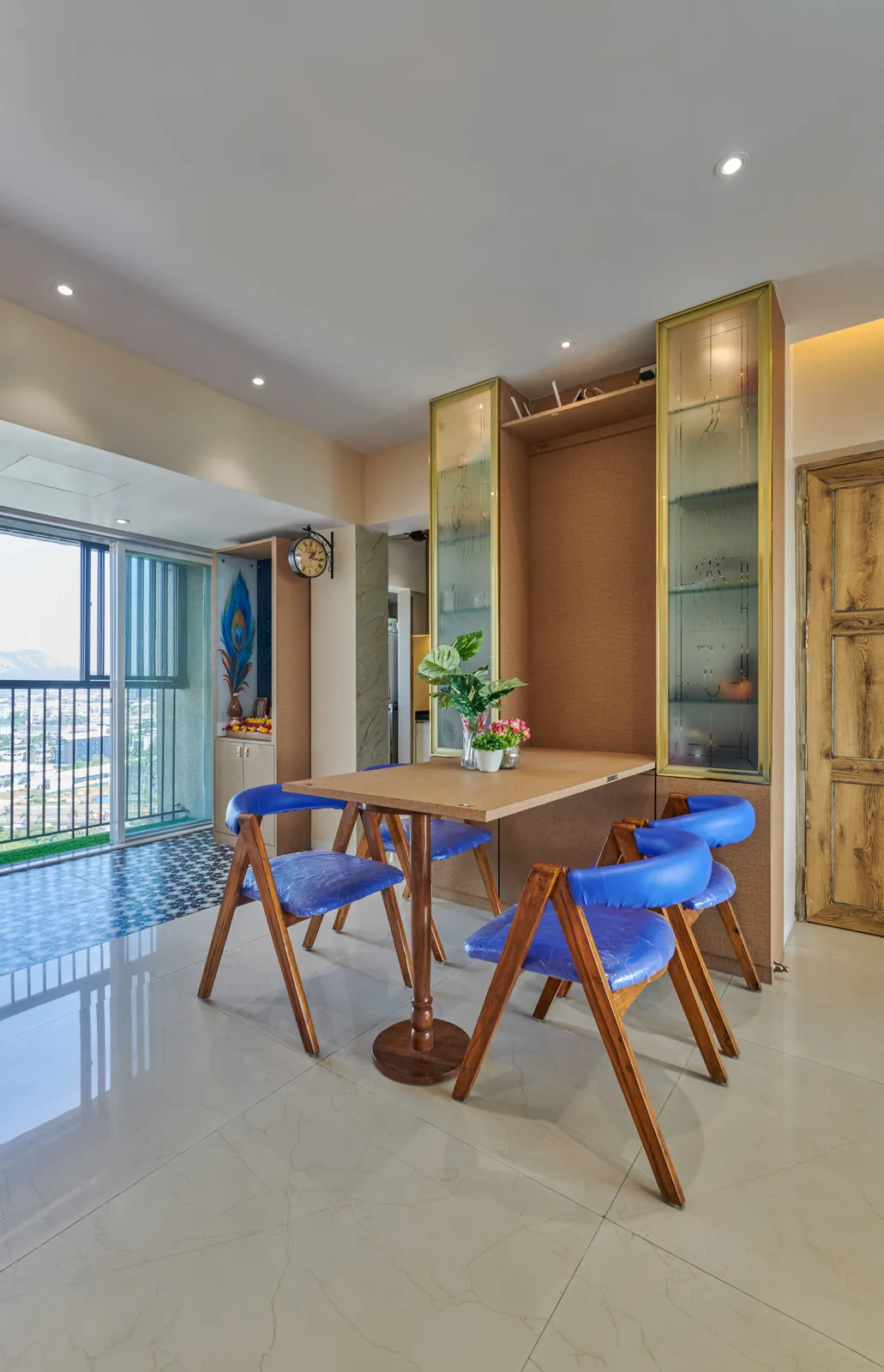 The compact dining area features a wooden table, blue upholstered chairs, and a frosted glass crockery unit, blending style and functionality.